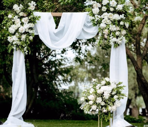 Elegant outdoor wedding ceremony setup at Copper Hills Golf Club in Oxford, Mi. featuring a white floral arch with greenery and draped white fabric, set against a serene backdrop.