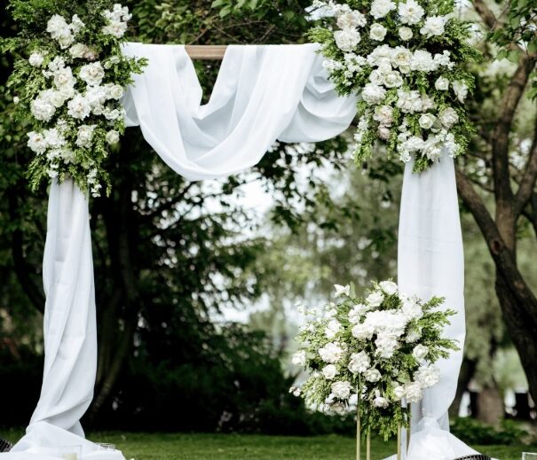 Elegant outdoor wedding ceremony setup at Copper Hills Golf Club in Oxford, Mi. featuring a white floral arch with greenery and draped white fabric, set against a serene backdrop.